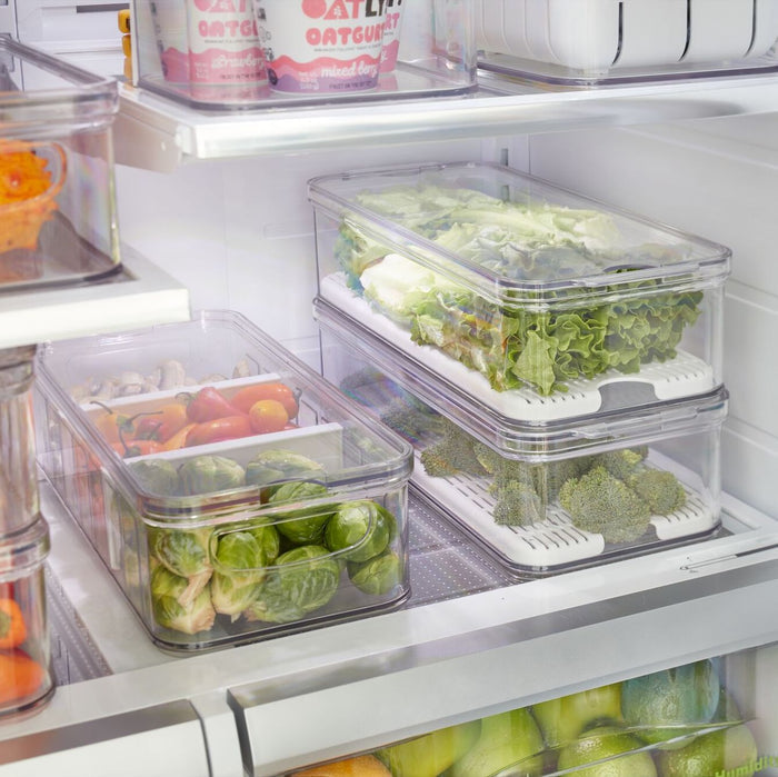 IDesigns Produce Fridge Bin, made with recycled plastic, showcases fresh vegetables like lettuce, broccoli, mushrooms, bell peppers, and Brussels sprouts. BPA-free containers are neatly arranged on refrigerator shelves with oat milk containers visible in the background.