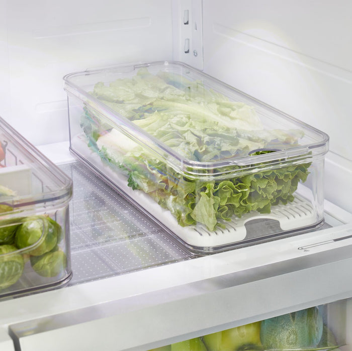 Inside the refrigerator, an iDesign Produce Fridge Bin made with recycled plastic and featuring integrated handles holds fresh lettuce. It sits neatly on a shelf surrounded by other faintly visible vegetables, ensuring an organized appearance.