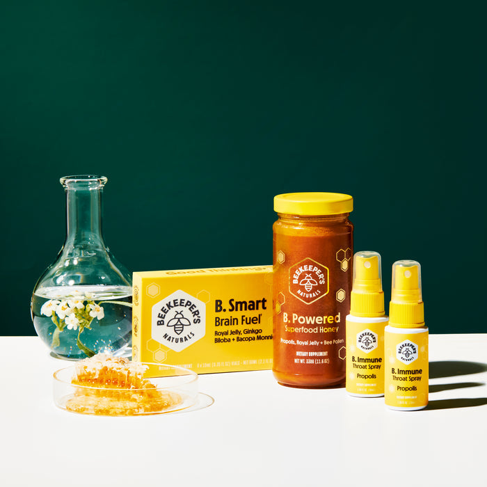 The Beekeepers Naturals collection features a jar of B. Powered Superfood Honey, a box of B. Smart Brain Fuel, two bottles of Propolis Immune Support Throat Spray, and a glass flask with flowers, with a honeycomb slice in the foreground.