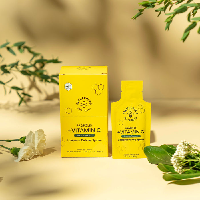 A beige surface with foliage features a Propolis Liposomal and Vitamin C package and sachet by Beekeepers Naturals, enhanced by bright lighting. The yellow design emphasizes immune support with a bee logo; shadows and a white flower add to the scene.