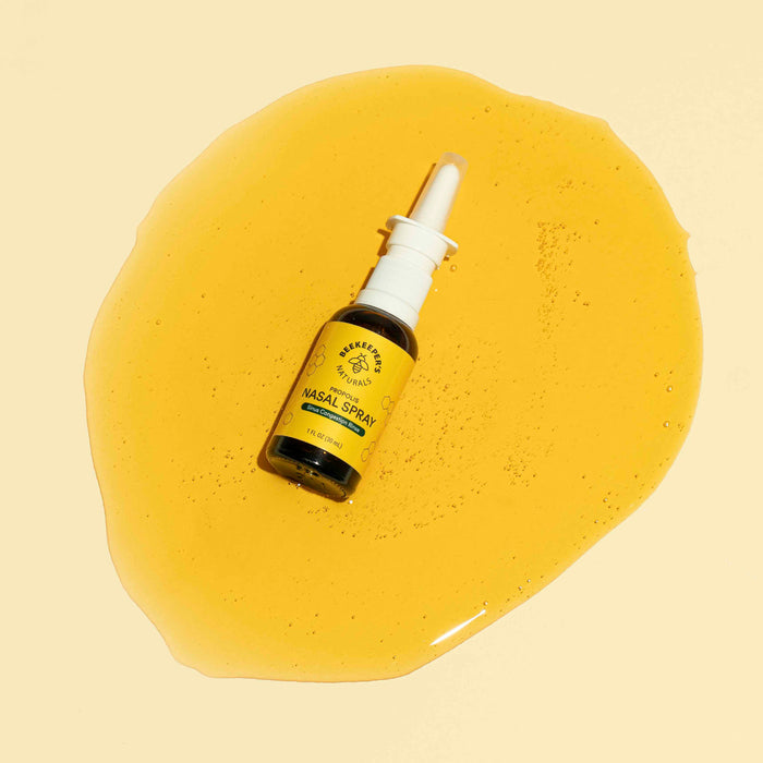 A Beekeepers Naturals Propolis Nasal Spray bottle sits in a pool of golden liquid, offering nasal relief. The backdrop is a solid, soft yellow.