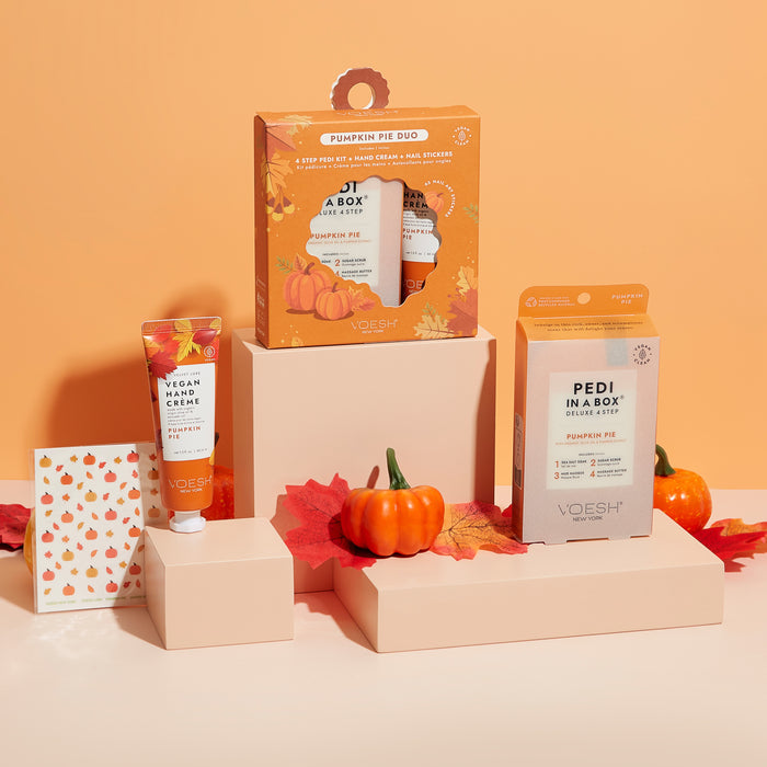 VOESH New York presents the Pumpkin Pie Duo with Nail Stickers, featuring a vegan body & hand crème and Pedi in a Box. Displayed on beige blocks against an orange backdrop, these skincare products celebrate autumn alongside decorative mini pumpkins and fall leaves.