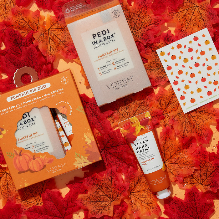 Flat lay of VOESH New Yorks Pumpkin Pie Duo with Nail Stickers set against red maple leaves, embodying autumn. Features a Pedi in a Box, vegan body & hand crème, and pumpkin nail stickers. Packaging showcases autumn colors and pumpkin designs.