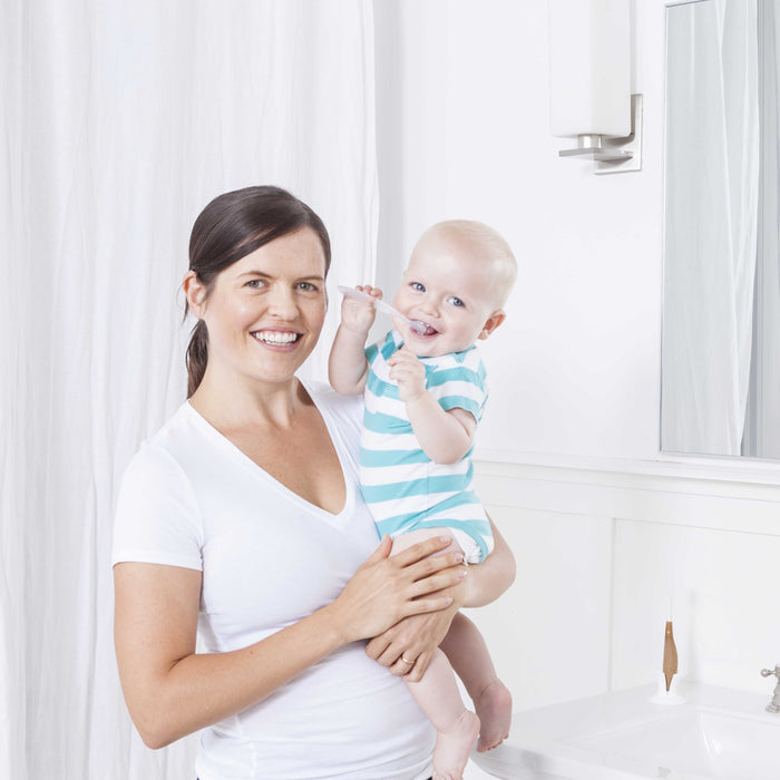 A smiling woman holds a baby in a striped onesie in a bright bathroom. The baby, holding the Radius Pure Brush 6 mo+ eco-friendly toothbrush, smiles near white curtains with a sink and mirror in the background.