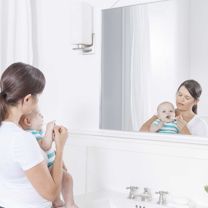 A woman with a baby in a striped onesie stands in a white bathroom, looking into the mirror. On the sink by shiny silver fixtures lies Radius Pure Brush 6 mo+, an eco-friendly toothbrush.
