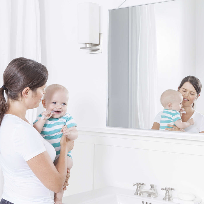 In a bathroom, a woman holds her baby, both facing the mirror. The baby in a striped onesie touches their mouth, highlighting the importance of choosing Radius Pure Brush 6 mo+ for gentle care of sensitive mouths.