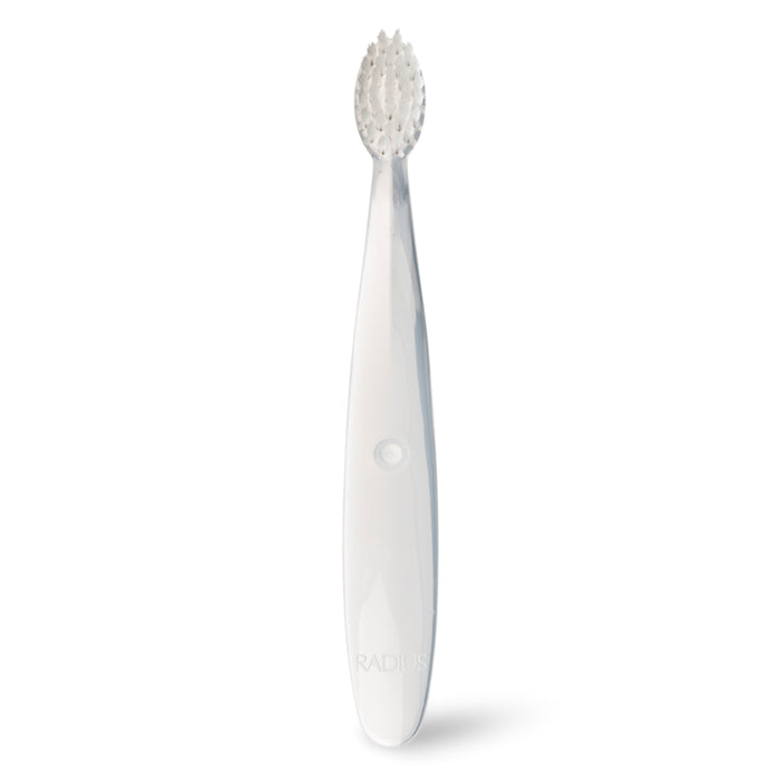 The Radius Pure Brush 6 mo+ is a white baby toothbrush with a wide handle and oval brush head featuring soft bristles for sensitive mouths, shown standing upright against a plain white background.