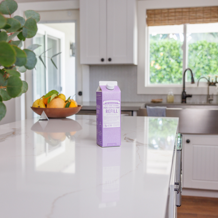 A Dr. Bronners Pure-Castile Liquid Soap Refill Carton with eco-friendly packaging in purple rests on a white marble kitchen countertop. In the background, a bowl of lemons and limes sits by a sink and large window showing lush greenery outside.