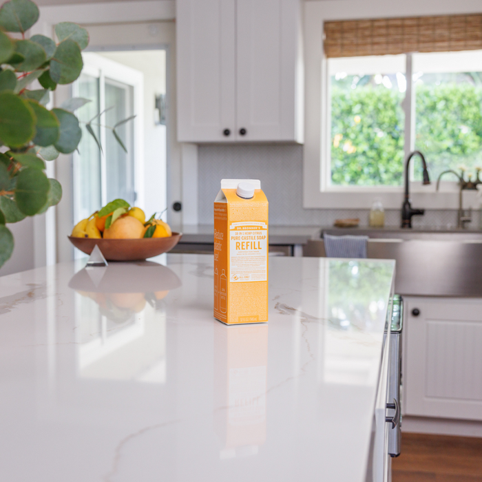 Dr. Bronners Pure-Castile Liquid Soap Refill Carton with eco-friendly packaging sits on a white marble kitchen island. A fruit bowl with limes and lemons is in the background beneath a large window above a farmhouse sink, with eucalyptus leaves partially visible on the left.
