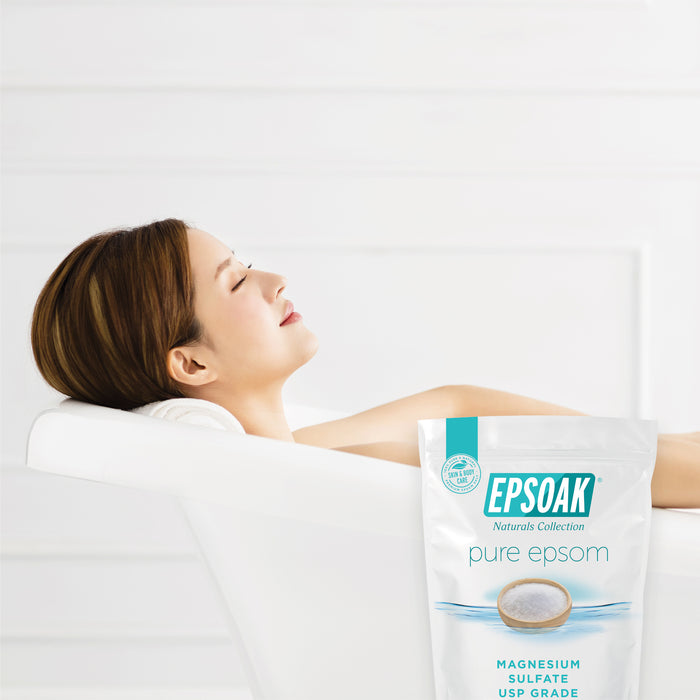 A woman relaxes in a white tub with her head on a towel, enjoying a soothing soak. In the foreground, a bag of San Francisco Salt Co Pure Epsom Salts - USP Grade promises muscle pain relief against a white background.