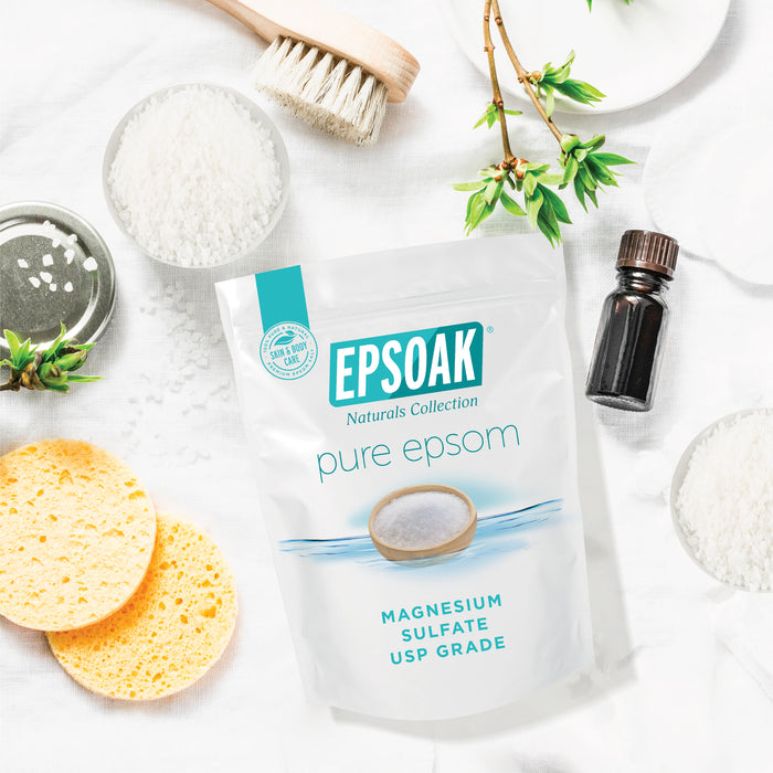 A bag of San Francisco Salt Co Pure Epsom Salts - USP Grade rests on a white surface with a yellow sponge, wooden brush, glass bottle, bowl of salt, leaves, and plate—ideal for soothing soaks and muscle relief.
