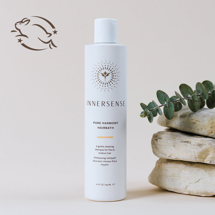 A bottle of Innersense Pure Harmony Hairbath is placed on a stone surface with eucalyptus sprigs. A logo featuring a leaping bunny is on the upper left, indicating a cruelty-free product. The background is a soft beige.