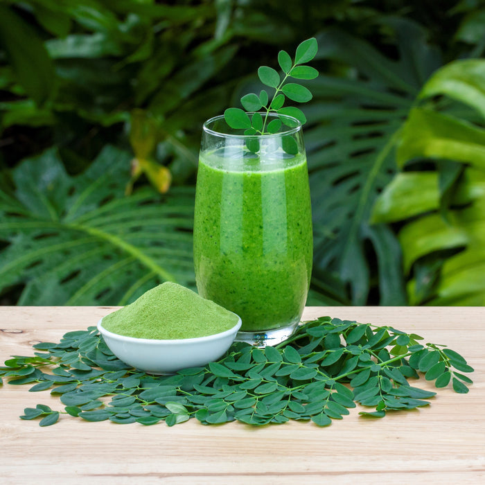 A glass of green smoothie garnished with a leafy sprig sits beside a bowl of Kuli Kuli Foods Pure Moringa Organic Superfood Powder on fresh leaves atop wood, with lush greenery in the background.