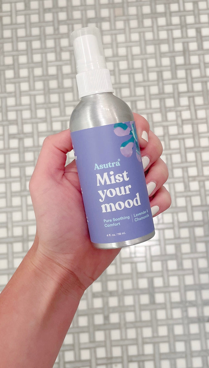 A hand holds a light purple bottle of Asutra Pure Soothing Comfort Aromatherapy Mist, featuring lavender and chamomile. Resting on a tiled surface, this vegan mist embodies tranquility to lift your mood.