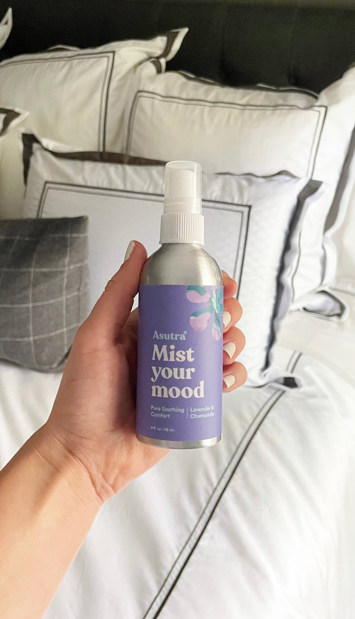 A hand holds a spray bottle labeled Pure Soothing Comfort Aromatherapy Mist from Asutra, featuring calming lavender and chamomile scents and a vegan formula. The backdrop displays a neatly made bed accented with white and gray pillows.