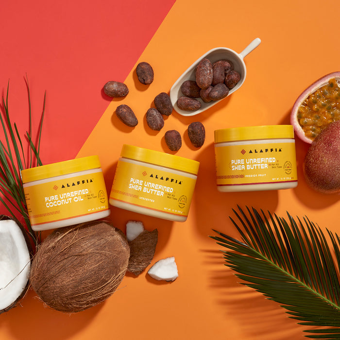 A colorful Alaffia skincare display features Pure Unrefined Shea Butter jars against a vivid orange and red backdrop, with nourishing coconut oil, whole and halved coconuts, shea nuts in a scoop, cracked passion fruit, and decorative palm leaves. Perfect for dry skin.