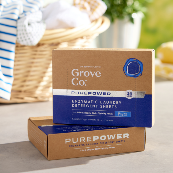 Two brown and blue boxes of Grove Co. PurePower Enzymatic Laundry Detergent Sheets labeled Sea Salt & Magnolia are stacked with a blurred laundry basket in the background. These bio-based products aim to reduce carbon emissions for a cleaner environment.