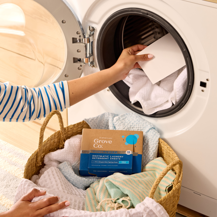 A person loads laundry into an open front-loading washer, holding a white PurePower Enzymatic Laundry Detergent Sheet. Next to the machine is a basket with towels and a box labeled Grove Co. PurePower Enzymatic Laundry Detergent Sheets, known for their bio-based cleansers reducing carbon emissions.