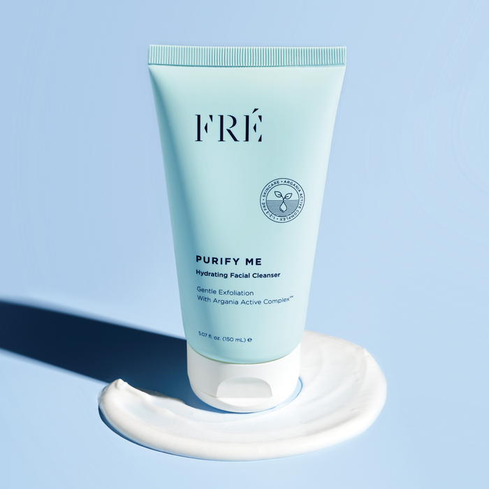 A 150 mL tube of FRÉ Skincares PURIFY ME Hydrating Facial Cleanser lies on a swirl of product against a blue backdrop, offering gentle exfoliation with Argania Active Complex and pore-minimizing benefits.