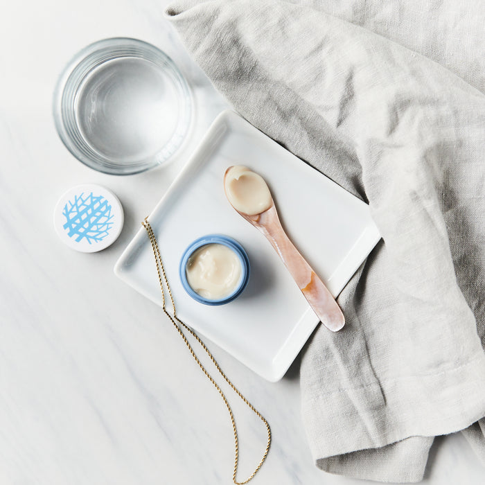 A small jar of Rooted Beautys Purifying De-puffing Eye Gel, rich in antioxidants, sits on a white square plate with a spoon beside a gold chain necklace. Nearby, a glass of water and white cloth rest on the marble surface, offering stylish relief for dark circles.