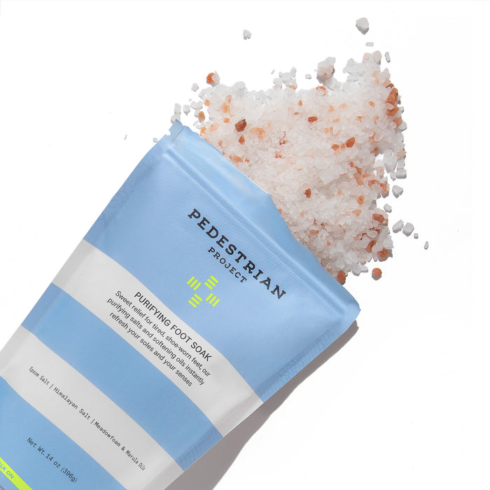 A Pedestrian Project packet labeled Purifying Foot Soak lies on its side, spilling coarse pink and white salt granules. The blue and white vegan packet is designed to detoxify your feet and weighs 7 oz (198g).