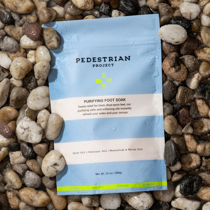 Pedestrian Projects Purifying Foot Soak, in a blue package glistening with water droplets, sits on wet pebbles. Its infused with vegan Epsom and Himalayan salts, plus meadowfoam and marula oils—ideal for rejuvenating tired feet.