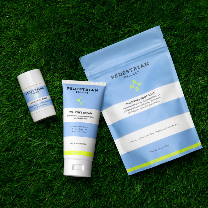 On the grass, Pedestrian Project products are elegantly displayed, featuring a cylindrical Dead Sea Heel Polish, a tube of Walkers Cream, and a detoxifying blue sachet of Purifying Foot Soak.