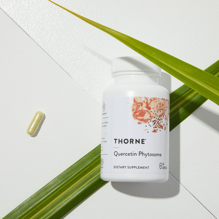 A Thorne Quercetin Phytosome bottle, recognized for antioxidant benefits, rests on geometric white surfaces with green palm leaves. A single capsule is nearby, and shadows form a minimalist scene.