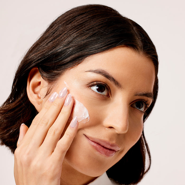 A person with dark hair gracefully applies the Acure Radically Rejuvenating Day Cream - SPF 30 to their cheek. Their gentle expression and manicured nails highlight their skincare routine against a plain, neutral background.