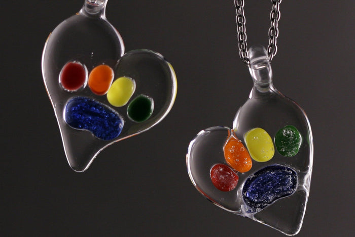 Two glass Rainbow Bridge pawprint pendants filled with bright colored beads on a dark background
