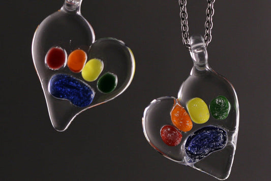 Two glass Rainbow Bridge pawprint pendants filled with bright colored beads on a dark background