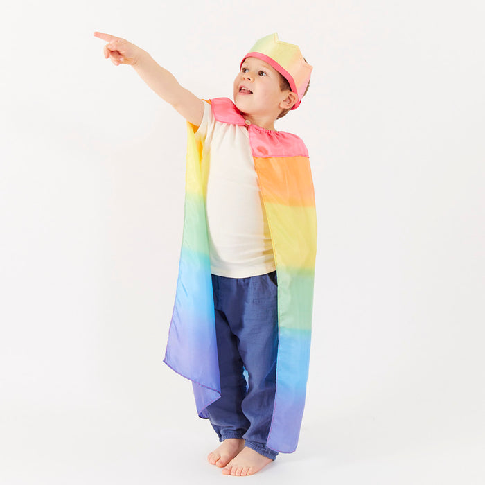 A child in a vibrant Rainbow Cape from Sarahs Silks and matching crown points left excitedly, barefoot in blue pants and a white shirt, embodying the joy of dramatic play against a plain white background.