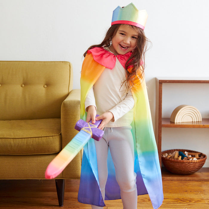 A child, immersed in open-ended play, dons Sarahs Silks Rainbow Cape and a colorful crown while holding a rainbow sword. They stand happily in the living room with a mustard-yellow armchair and a wooden shelf as their backdrop.