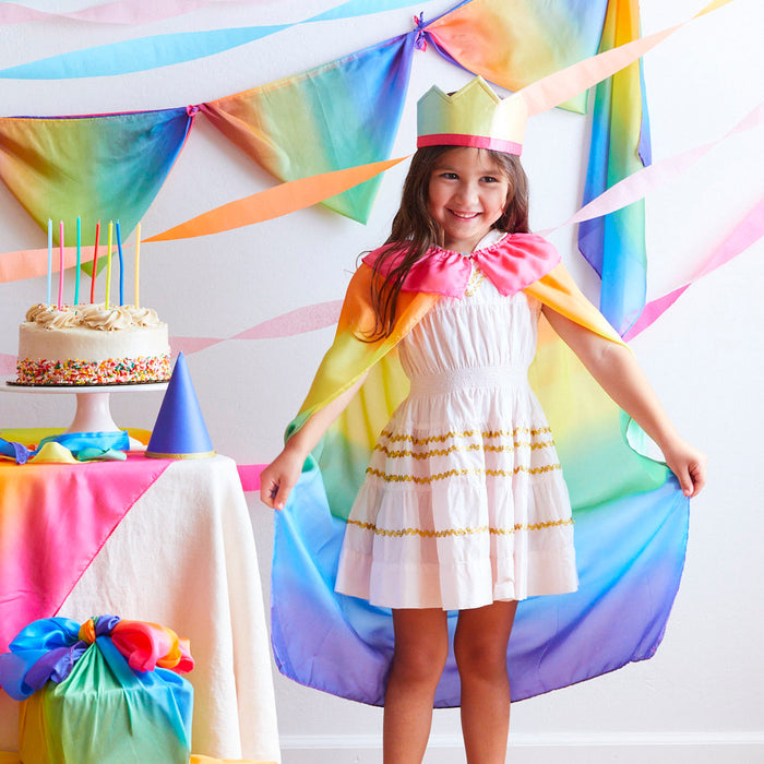 A child in a white dress and Sarahs Silks Rainbow Cape smiles at a rainbow-themed birthday party, embodying the spirit of dress-up play. A table with a rainbow cake and colorful decorations adds to the whimsical scene in the background.