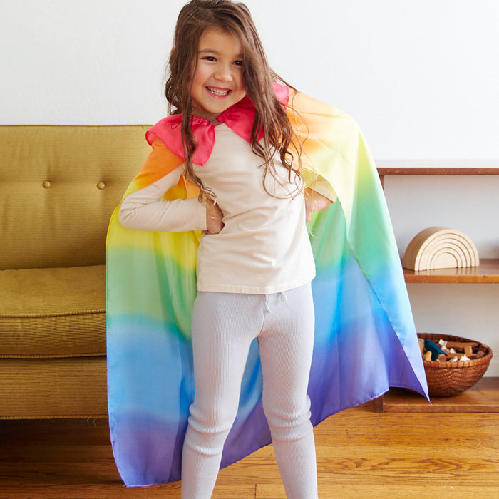 A child with long hair smiles, posing in a colorful Rainbow Cape from Sarahs Silks and a pink collar. Captivated by dress-up magic, they stand indoors near a couch and shelf decorated with small wooden items.