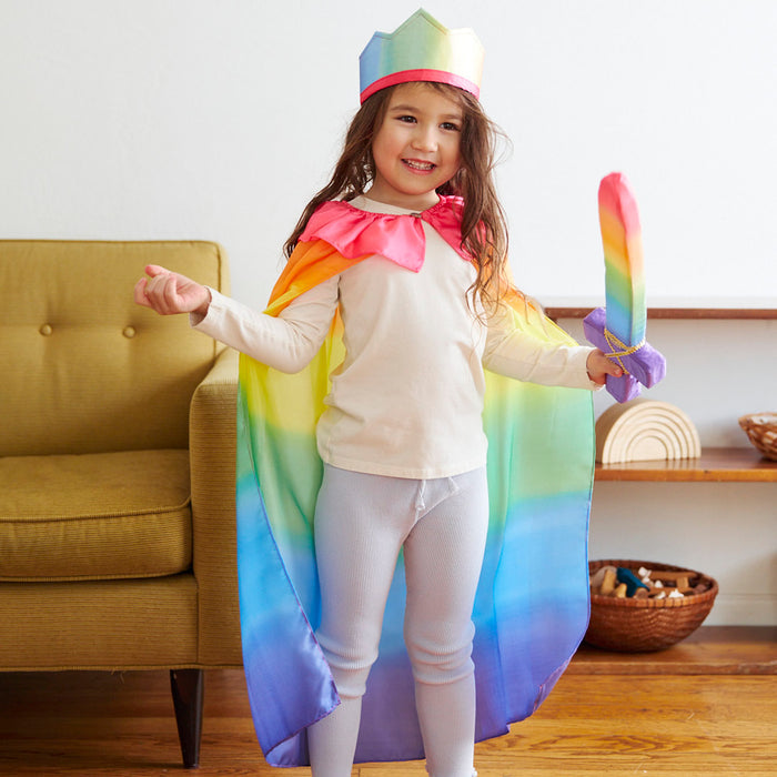 A child beams while engaged in dress-up play, donning a vibrant crown, Sarahs Silks Rainbow Cape flowing elegantly on their shoulders, and a matching rainbow sword in hand. Their long hair flows as they proudly stand near a yellow chair and basket.