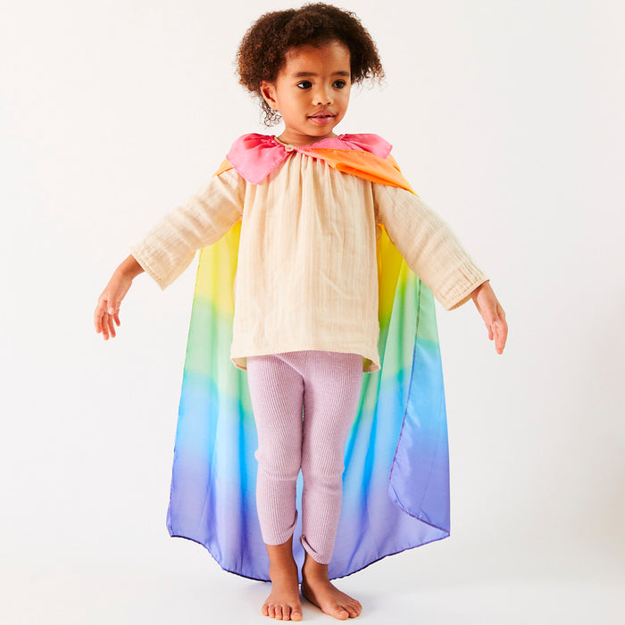 A child in a cream top and pink leggings poses with arms outstretched, wearing the vibrant Rainbow Cape by Sarahs Silks with a pink collar. Set against a plain white background, its perfect for sparking imagination in dress-up play.