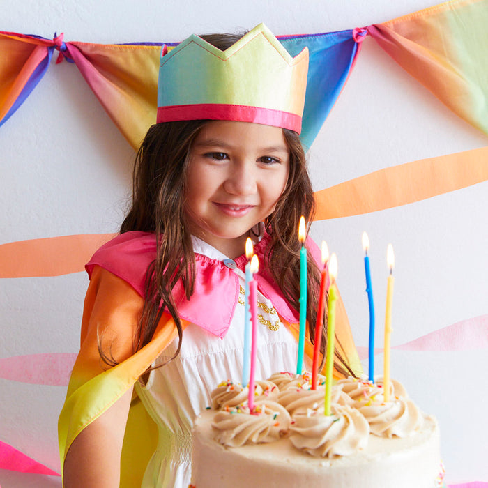 A young girl in a Sarahs Silks Rainbow Crown and cape smiles behind a candlelit cake, with a rainbow garland in the background, setting a festive scene for imaginative play.