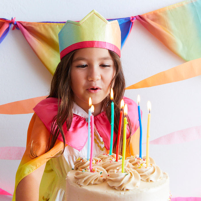 A child, lost in imaginative play, dons a Rainbow Crown from Sarahs Silks and a silk cape while blowing out birthday candles. The cake, adorned with beige frosting and several lit candles, is surrounded by vibrant streamers for a festive touch.
