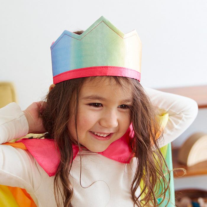 A child wears a Rainbow Crown from Sarahs Silks, embracing imaginative play. The vibrant crown adds magic as they smile and relax with their hands behind their head, while the rainbow-adorned cape contrasts beautifully against the softly lit room.