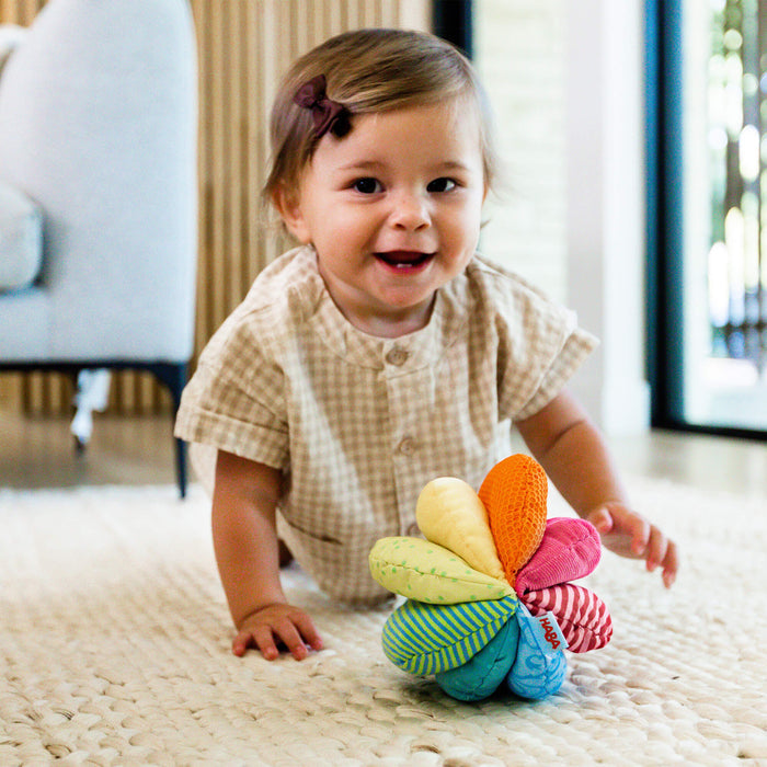 A smiling toddler in a beige checkered outfit crawls on a light rug towards a HABA Rainbow Fabric Baby Ball with 8 Different Sensory Effects, enhancing motor skills. A white chair and wooden wall panels create a cozy background.