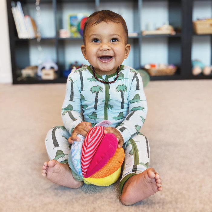 A smiling baby sits on a carpeted floor in a palm tree onesie, holding a HABA Rainbow Fabric Baby Ball with 8 Different Sensory Effects for sensory play and motor skill development. A bookshelf of toys and books is blurred in the background.