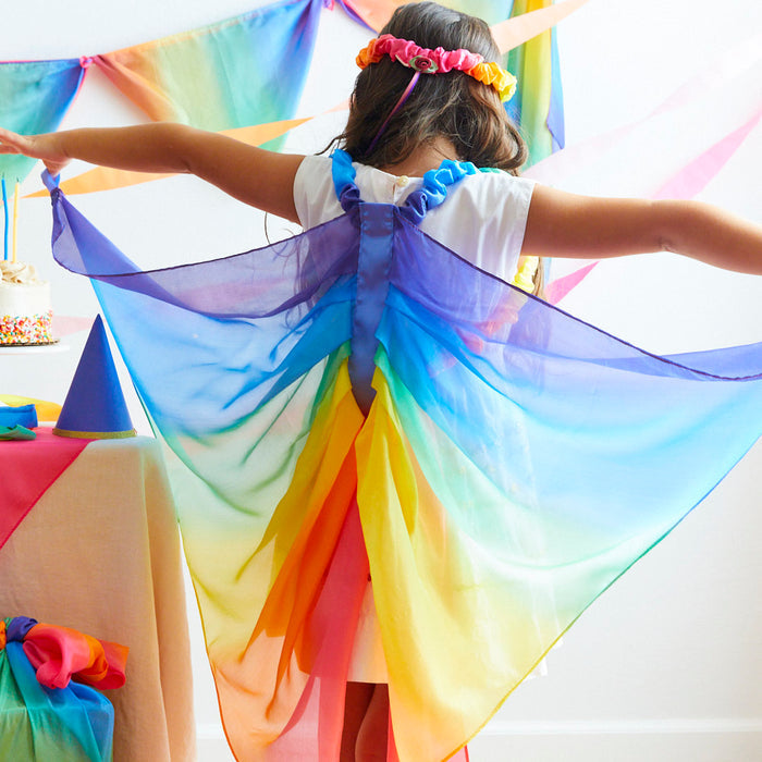 A child with Rainbow Fairy Wings from Sarahs Silks and a flower crown stands with arms outstretched beside a table adorned with colorful decorations, a cone hat, and a cake. Streamers hang in the background, crafting an ideal dress-up play scene.