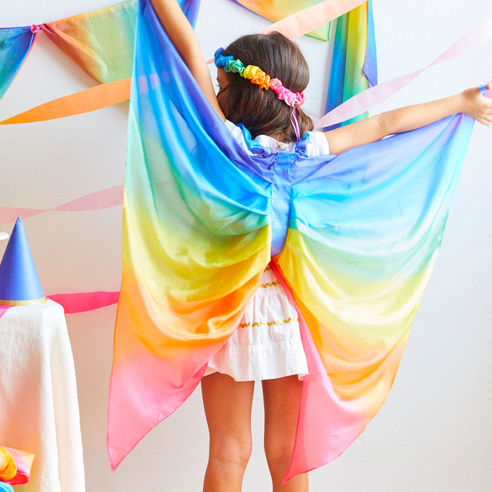 A child in Sarahs Silks Rainbow Fairy Wings stands with arms outstretched, wearing a white mulberry silk outfit and a flower crown, amidst festive decorations.