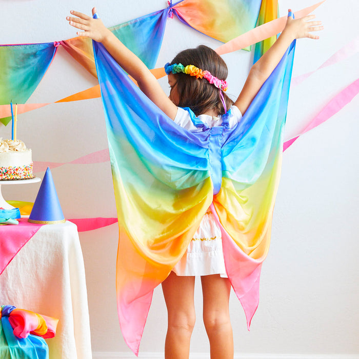 A child in a white dress and Sarahs Silks Rainbow Fairy Wings stands with arms outstretched, embodying the joy of dress-up play. The room features rainbow-themed decorations, with banners and a cake. A flower crown on their head adds magic to the air.