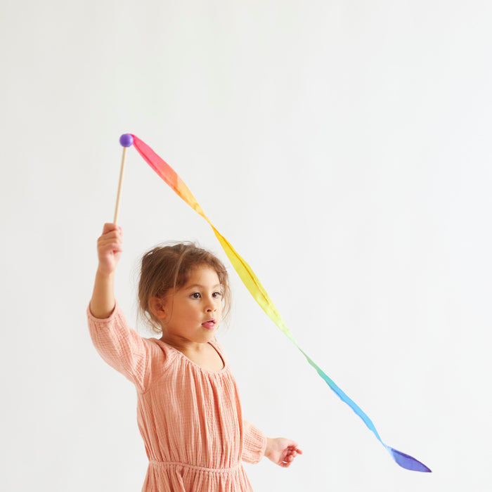 A young girl in a pink dress joyfully twirls Sarahs Silks Rainbow Streamer, embracing open-ended play against a plain white background.