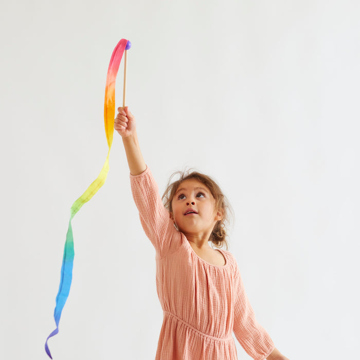 A young girl in a peach dress joyfully waves Sarahs Silks Rainbow Streamer against a plain background, her expression focused and curious as she engages in open-ended play.