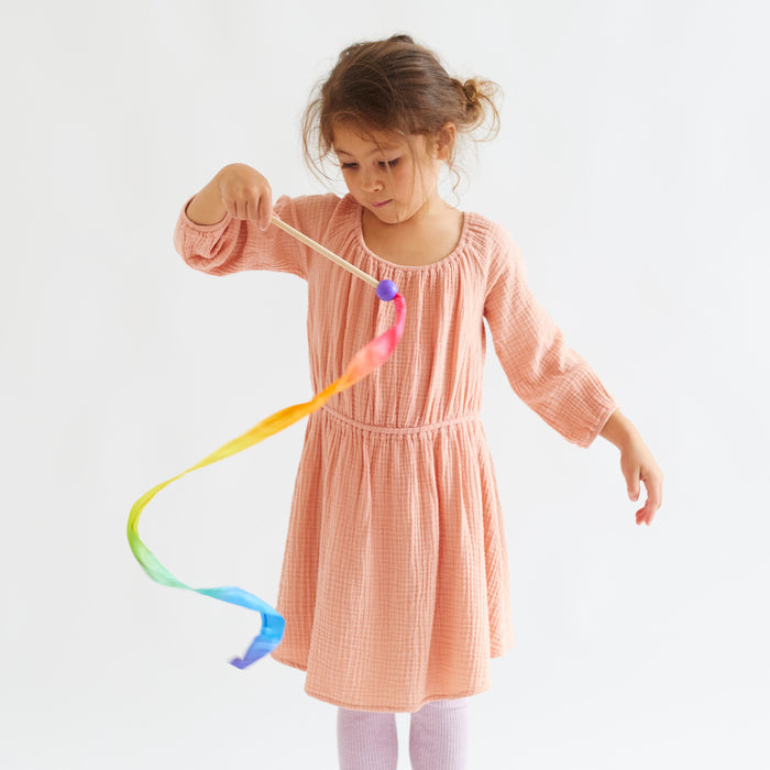 A young girl in a peach dress joyfully plays with her Rainbow Streamer by Sarahs Silks. The colorful ribbon swirls gracefully around her, creating mesmerizing patterns against the plain white background.
