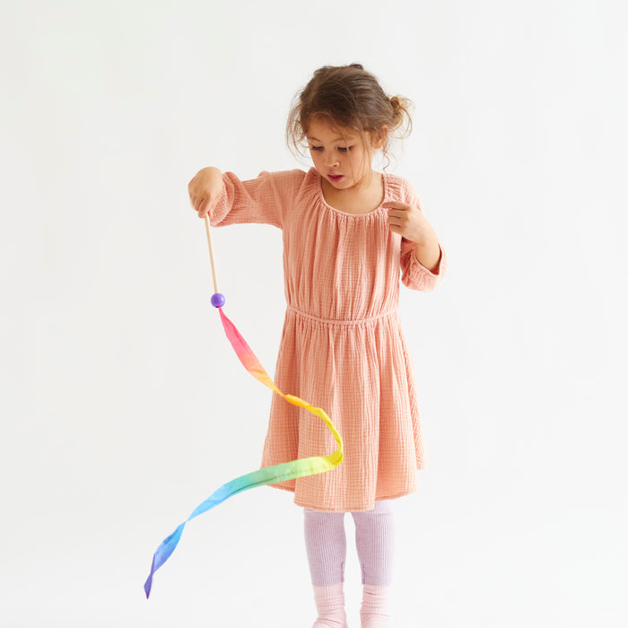A young girl, dressed in peach, twirls the Rainbow Streamer from Sarahs Silks against a white backdrop. Focused and engaged, she grips the vibrant silk wand with both hands, embodying the magic of open-ended play.