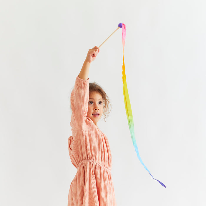 A joyful child in a pink dress waves Sarahs Silks Rainbow Streamer, displaying its colorful gradient against a plain white background and capturing the essence of open-ended play.